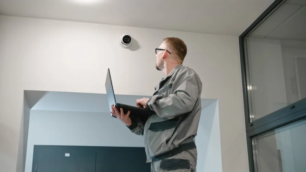Installer in uniform sets up security camera in office room using laptop. Man in glasses checks CCTV cameras in computer program. Monitoring system. Concept of surveillance system and privacy.